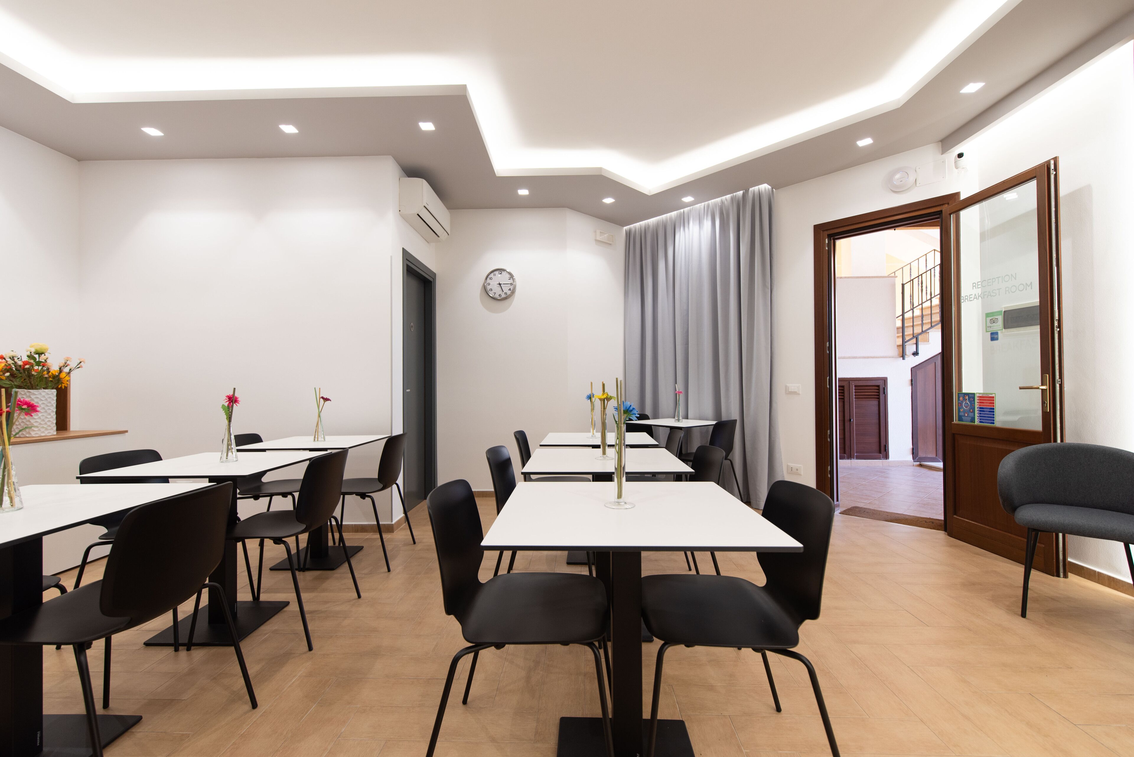 A hotel dining area with multiple tables and chairs, a clock on the wall, and a staircase leading to another floor.