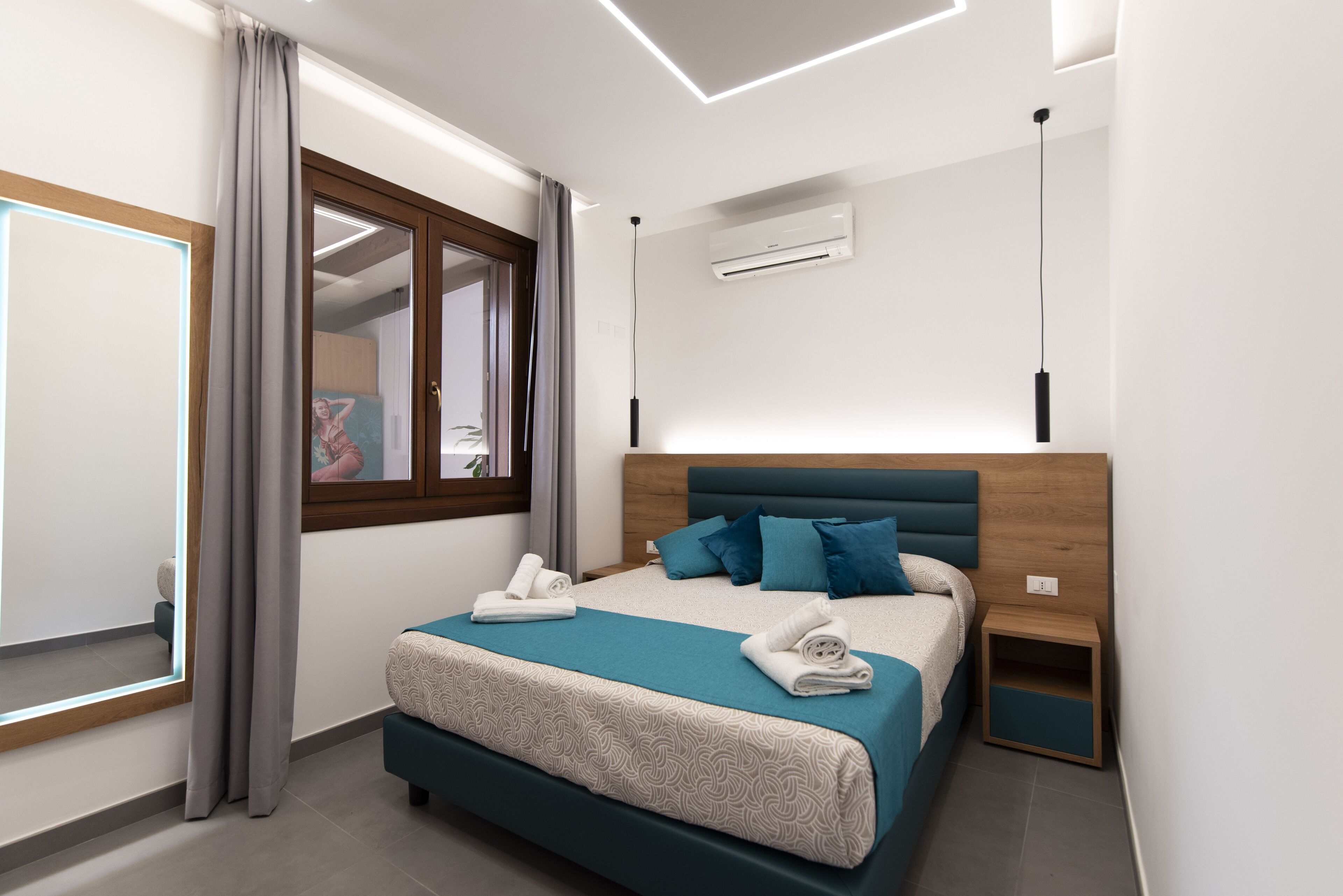 A modern bedroom with a bed, bedside tables, a window with curtains, an air conditioning unit, and a mirror.