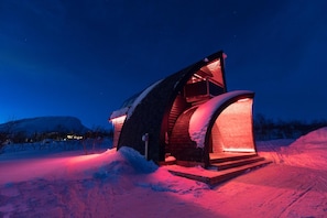 Un edificio moderno de forma arqueada, iluminado cálidamente desde el interior y rodeado de nieve.