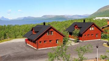 Two red wooden cabins with black roofs, a paved parking area, and a view of a lake and mountains in the background.