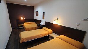 A hotel room with two single beds, a small table, a chair, and a wall-mounted lamp.