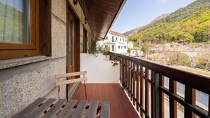 Un balcon avec un banc en bois, un bâtiment blanc avec des volets verts en arrière-plan, et un paysage montagneux.