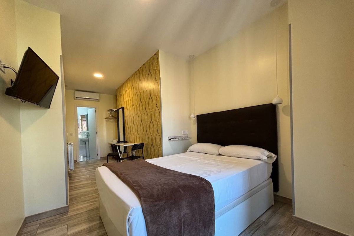 A hotel room with a bed, a flat-screen TV mounted on the wall, a small table with a chair, and a wooden floor.