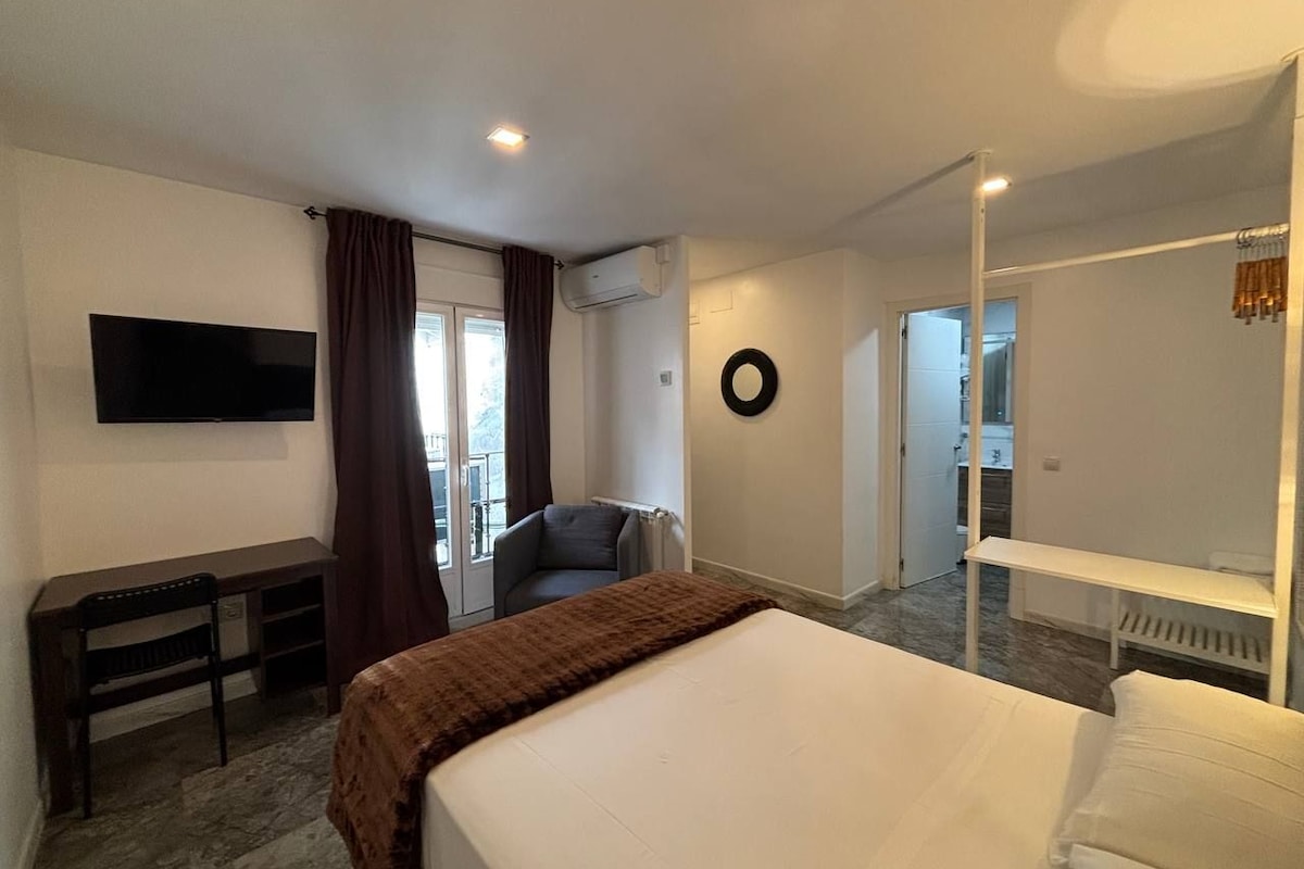 A hotel room with a bed, a desk, a chair, a TV, and a view of the outside.