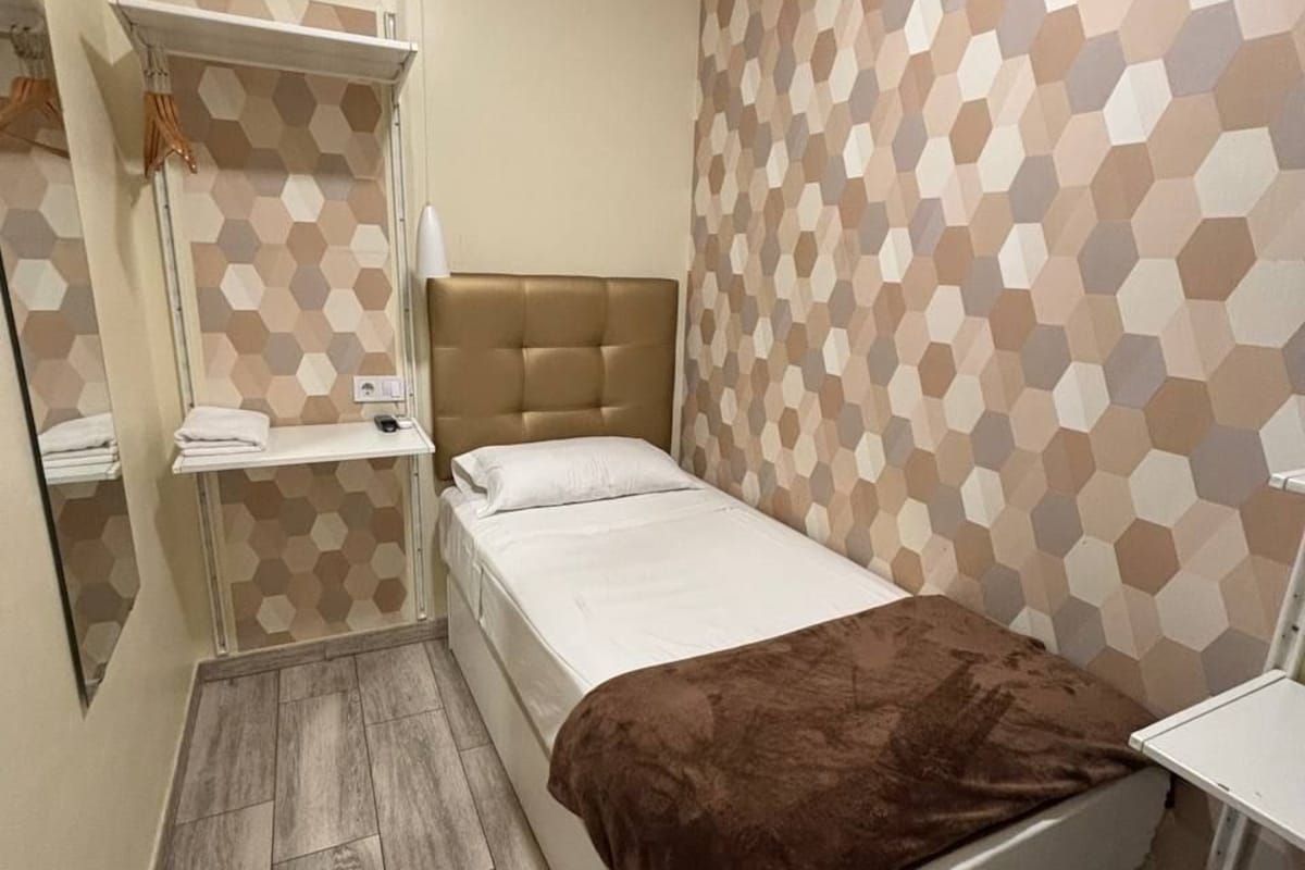 A single bed with a brown blanket, a bedside table, a mirror, and a wall with a geometric pattern.