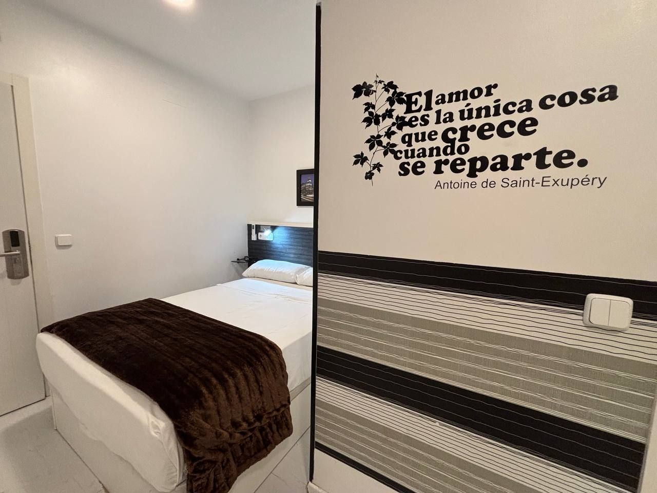 A small, modern bedroom with a bed, a bedside table, and a wall with a decorative quote.