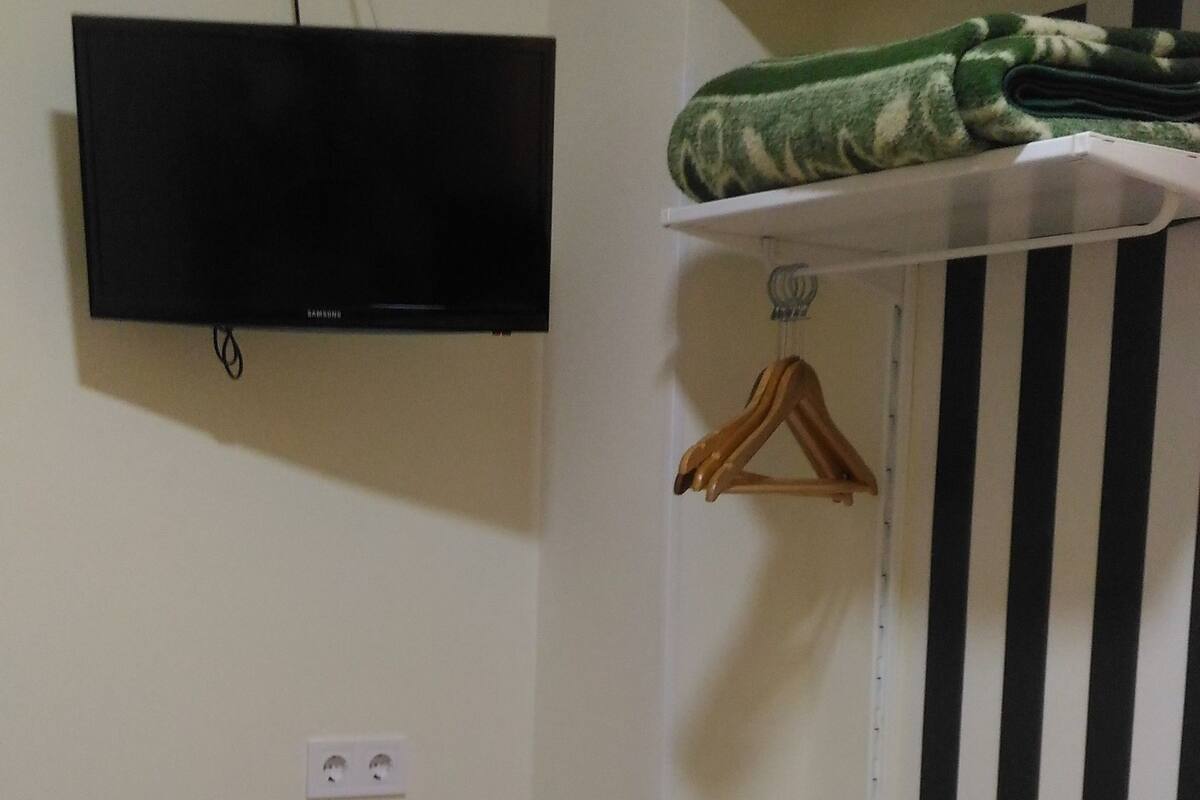 A room with a bed, a TV mounted on the wall, a radiator, a striped curtain, and a shelf with folded towels.