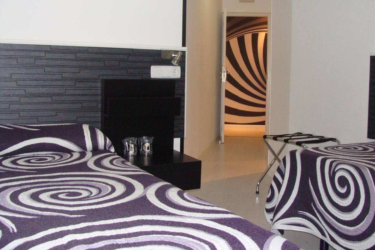 A hotel room with a modern design, featuring a black headboard, a small bedside table with a lamp, and a patterned bedspread.