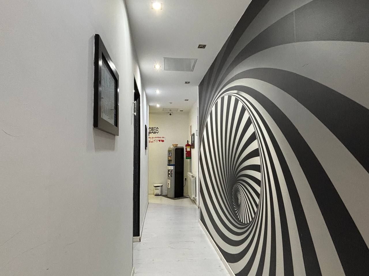 Un pasillo con diseño de rayas negras y blancas en la pared, un cuadro enmarcado a la izquierda y una máquina expendedora al fondo.