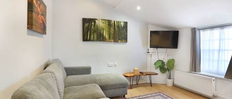 A modern living room with a grey sectional sofa, a large forest-themed painting, a flat-screen TV, a small wooden side table, and a potted plant.