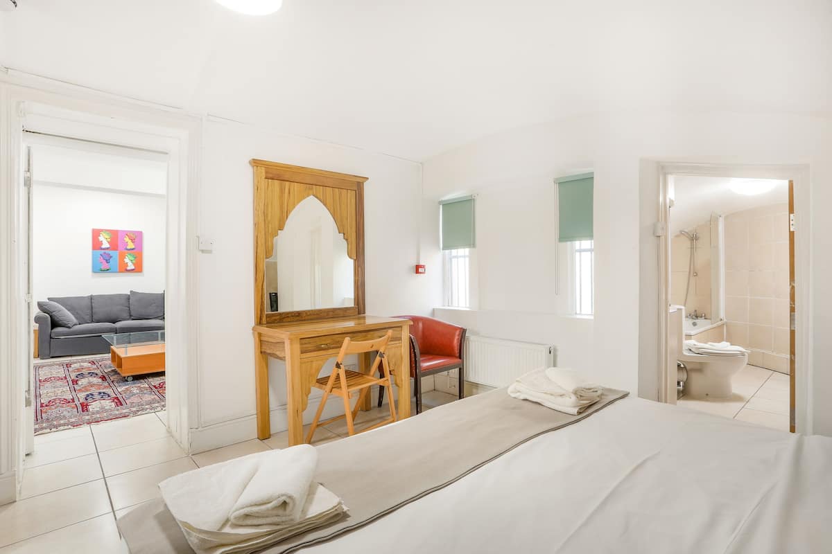 Een slaapkamer met een bed, een houten bureau, een stoel, een rode stoel, een spiegel, een raam en een badkamer die door een geopende deur zichtbaar is.