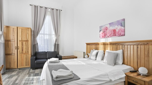 A hotel room with a bed, a wooden headboard, a grey sofa, a wooden wardrobe, and a painting of roses on the wall. - BeiRoom Hyde Park Apartments (London)