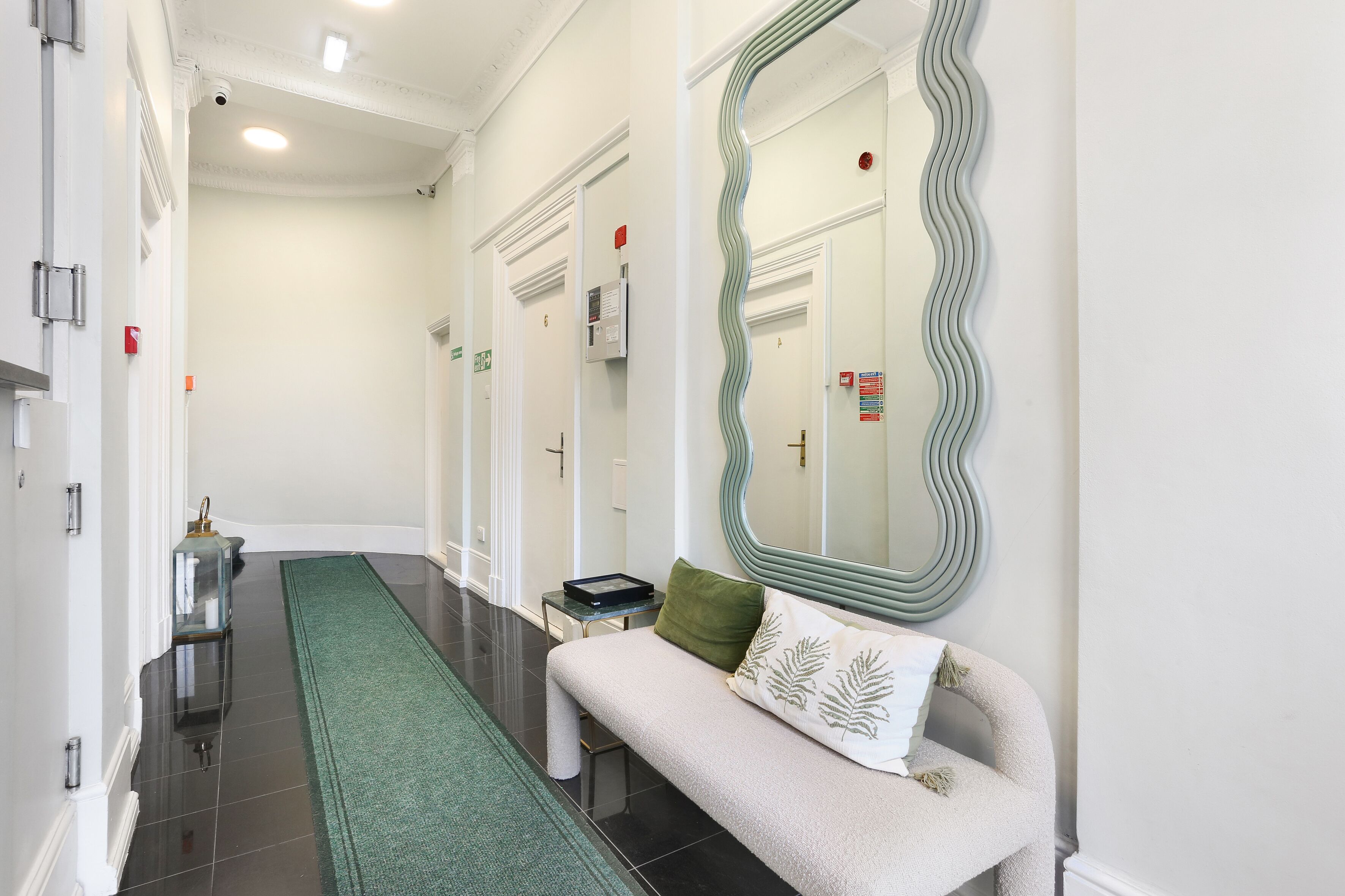 Un couloir avec une moquette verte, un banc avec un coussin vert, un grand miroir et une porte dotée d’une lumière rouge d’urgence.