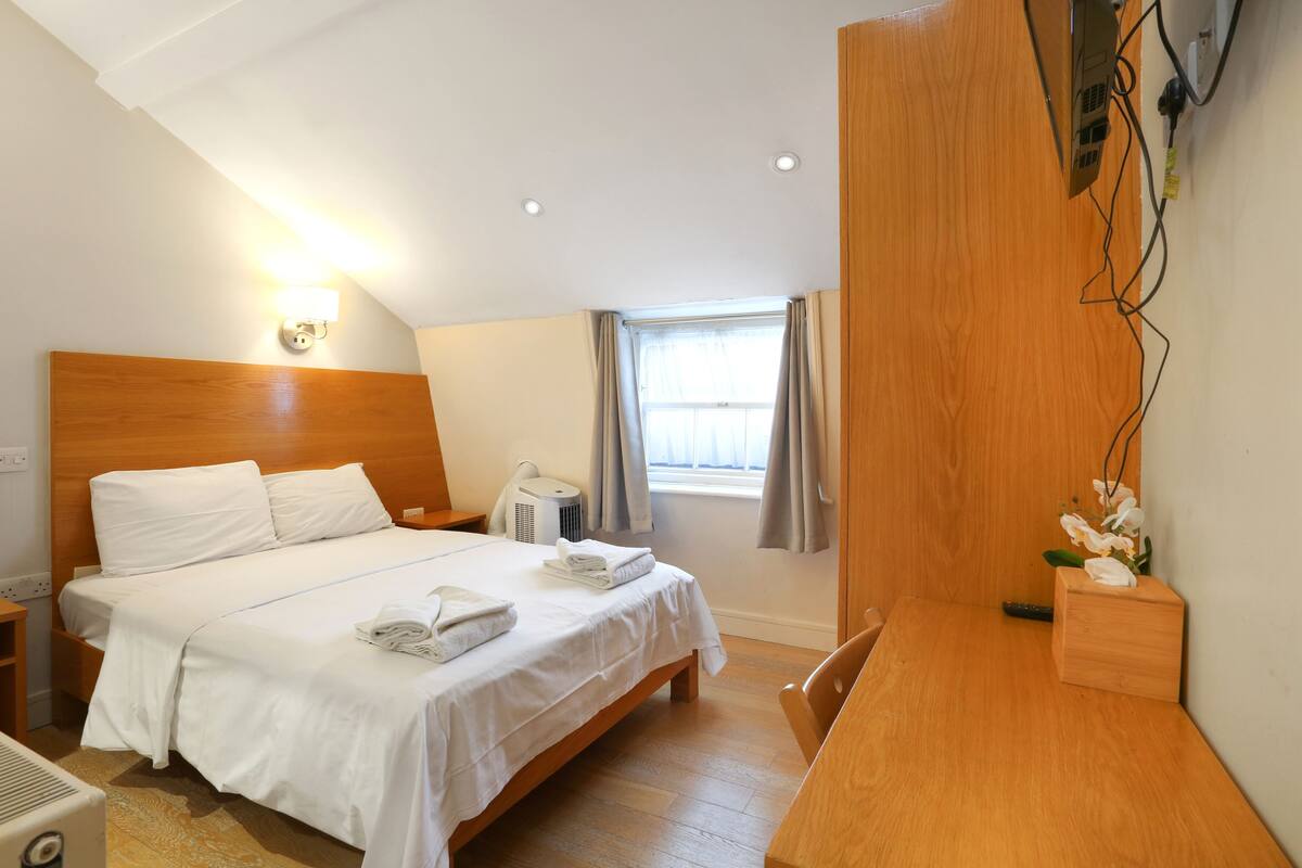 Hotelkamer met houten hoofdeinde, bed met witte lakens, nachtkastje, bureau, stoel, raam met gordijnen en een wandgemonteerde airconditioner.
