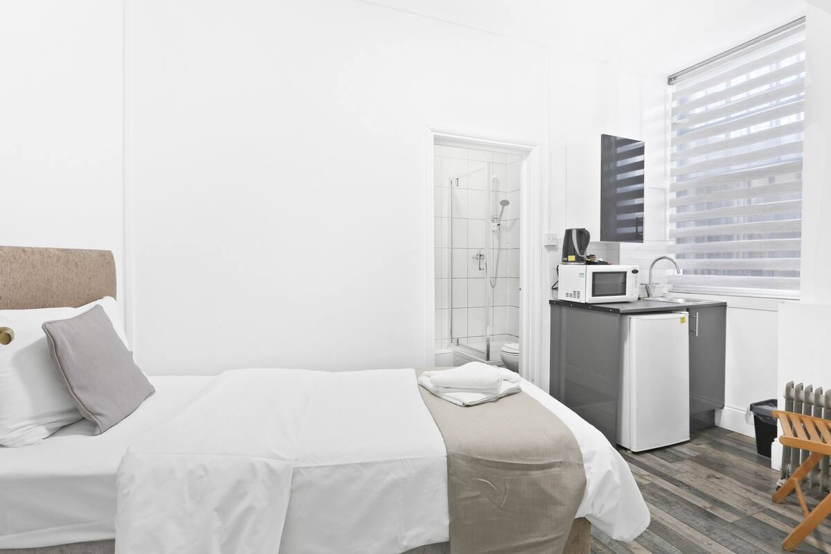 Een hotelkamer met een bed, een kleine kitchenette, een douche en een raam met jaloezieën.