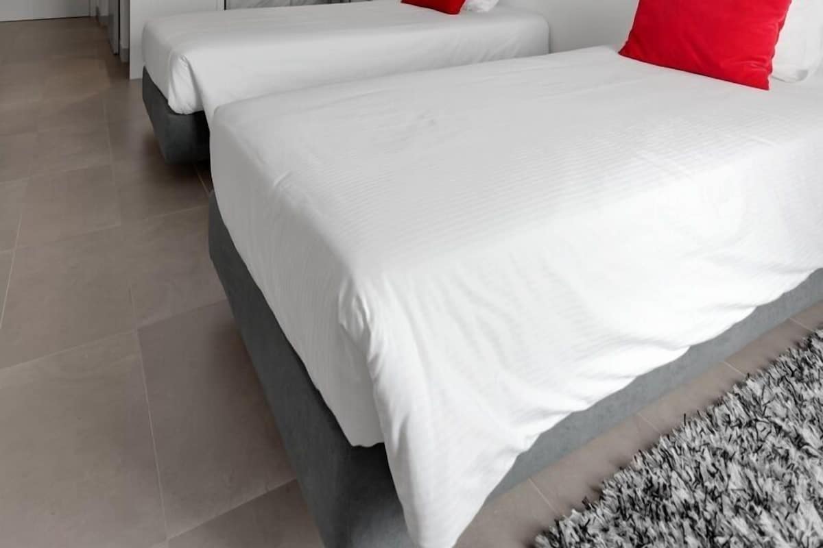 Una habitación de hotel moderna con dos camas, cabecero gris y almohadones rojos.