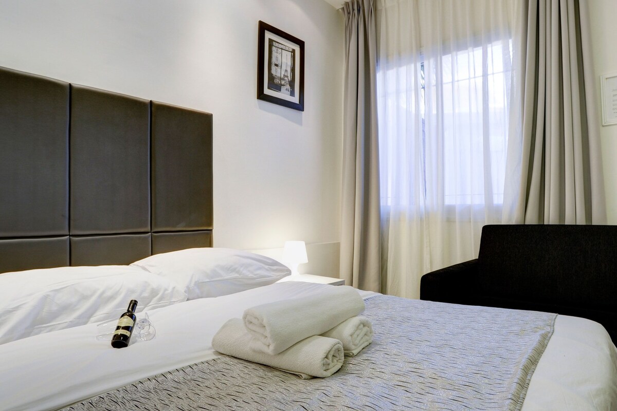 Una cama bien hecha con una botella y toallas, un cabecero, un cuadro enmarco y una ventana con cortinas.