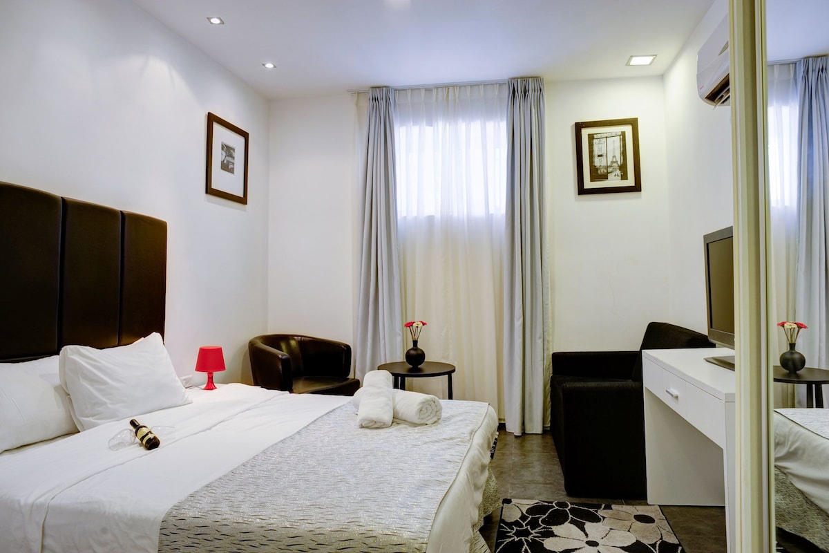 Habitación de hotel con cama, escritorio, silla, una mesita de noche con un jarrón y espejo.