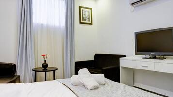 Een hotelkamer met een bed, een televisie op een standaard, een stoel, een klein tafeltje met een vaas, en een foto aan de muur.