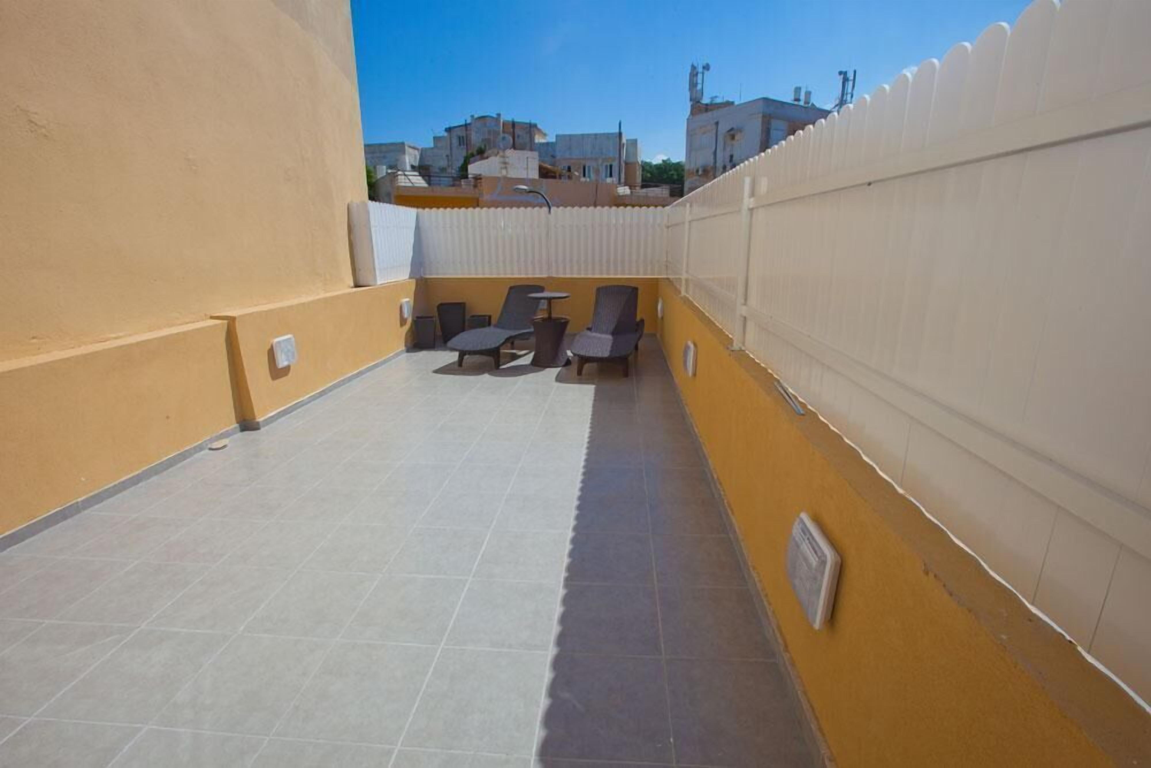 Una terraza en la azotea con zona de estar, valla blanca y vista de edificios al fondo.