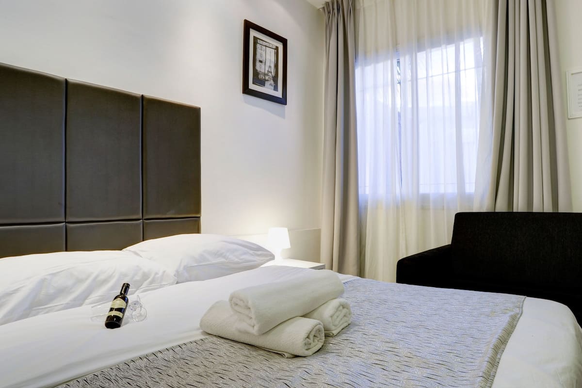 A neatly made bed with a bottle and towels, a headboard, a framed picture, and a window with curtains.
