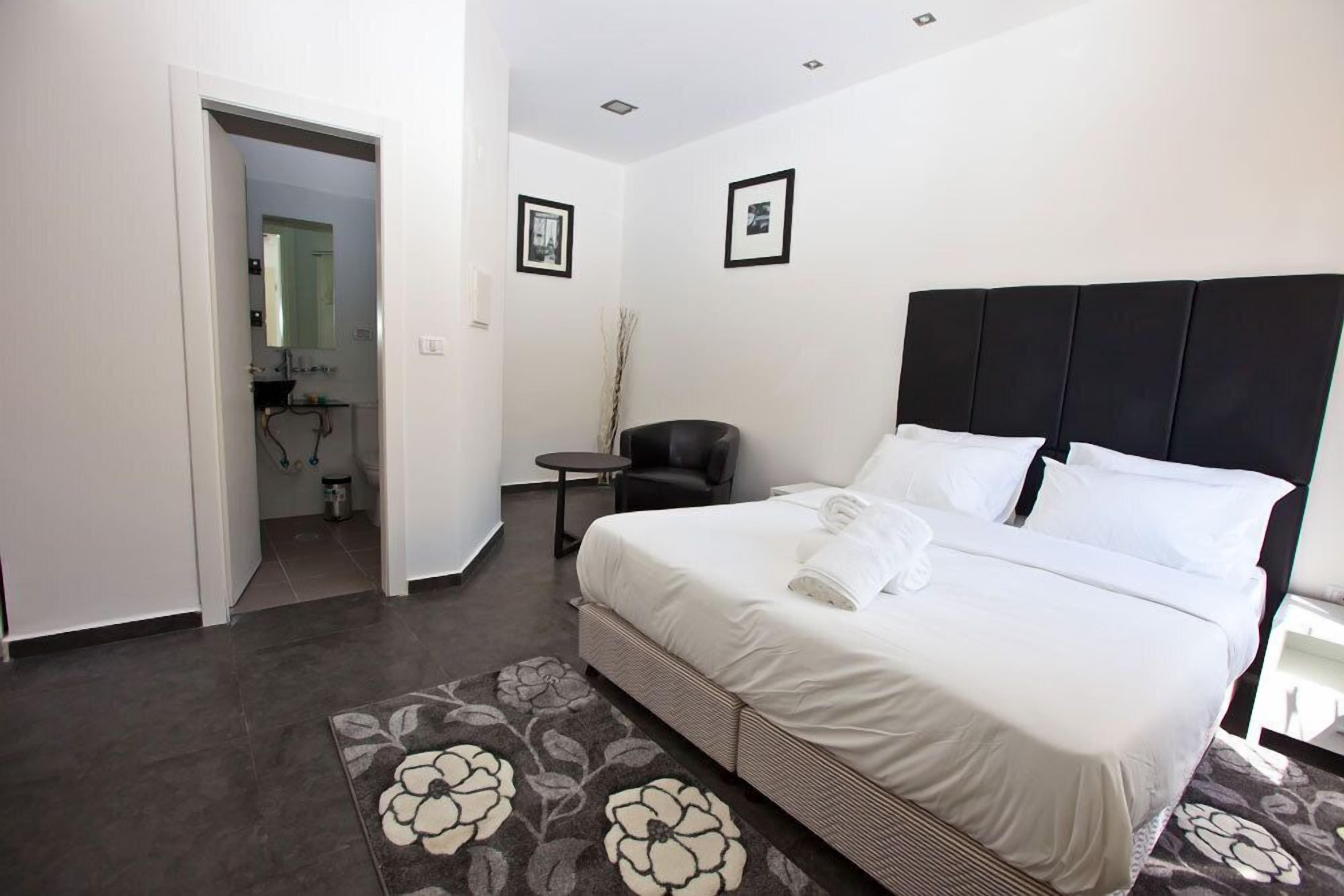 Une chambre à coucher avec un grand lit, une petite table, une chaise et une salle de bain visible à travers une porte ouverte.