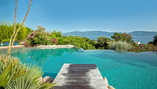 A clear blue infinity pool with a wooden deck, surrounded by lush greenery and a view of the sea and mountains. - Résidence Arco Plage (Olmeto)