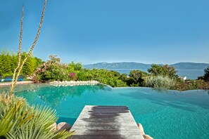 A clear blue infinity pool with a wooden deck, surrounded by lush greenery and a view of the sea and mountains. - Résidence Arco Plage (Olmeto)