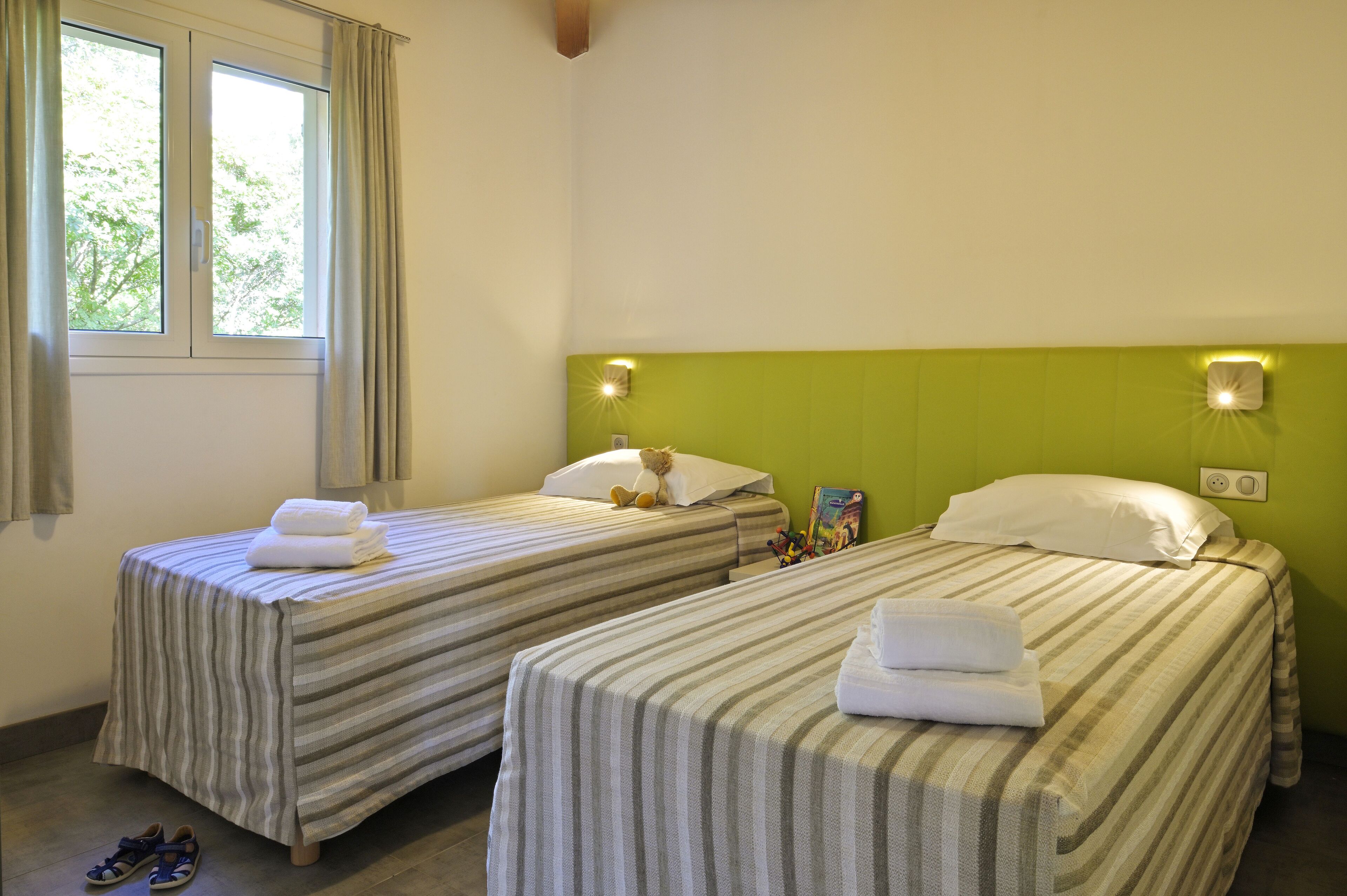 Habitación de hotel con dos camas individuales, cada una con colcha a rayas y una almohada. Hay toallas sobre las camas y un peluche sobre una de ellas. La habitación tiene un cabecero verde y una ventana con cortinas.