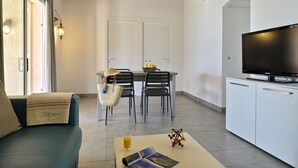 A modern living room with a sofa, a coffee table with a magazine and a glass of orange juice, a TV, and a dining area with chairs and a table. - Résidence Arco Plage (Olmeto)