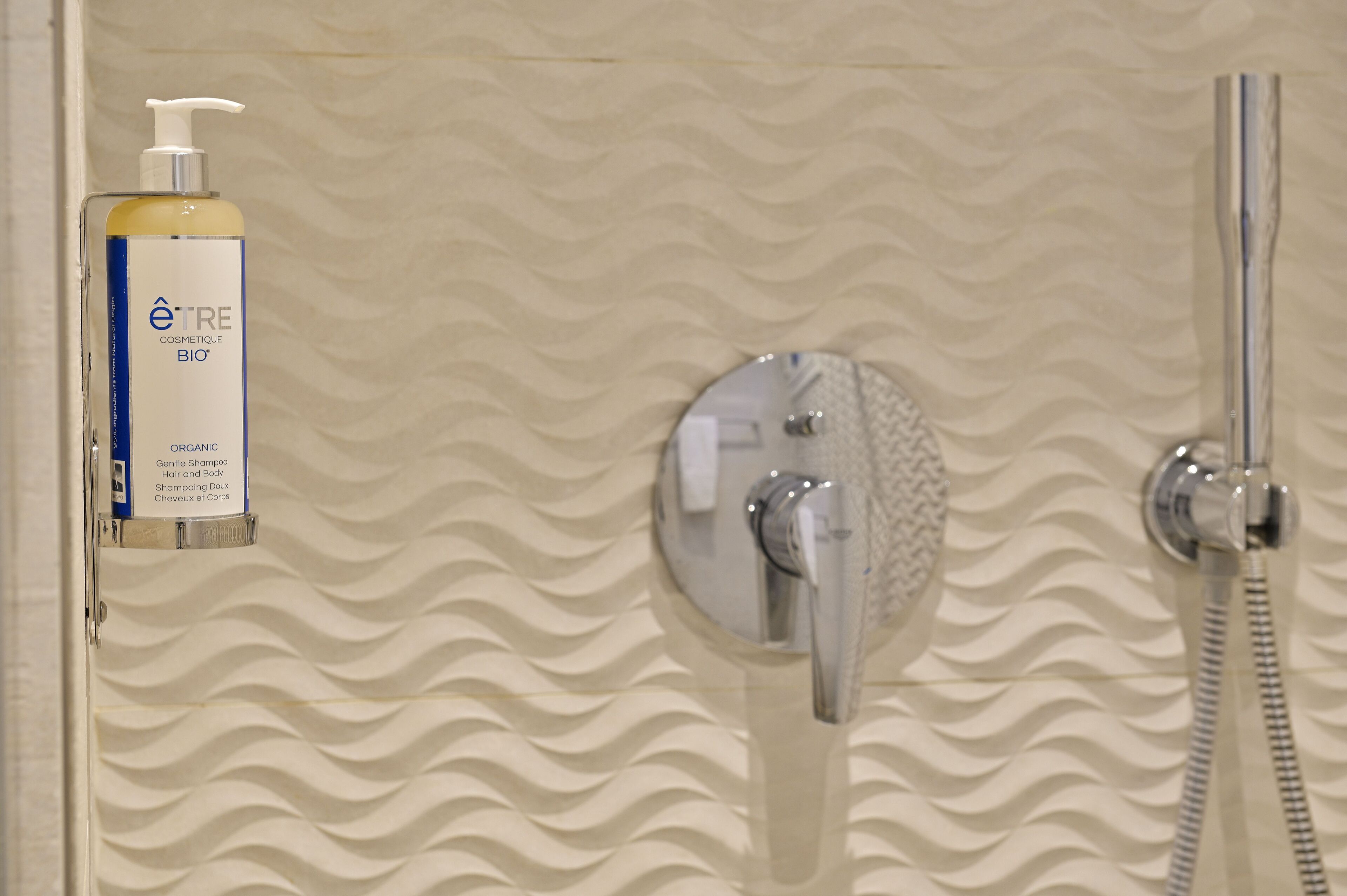 Un baño con pared texturizada, ducha con cabezal de ducha manual y una botella de champú orgánico.