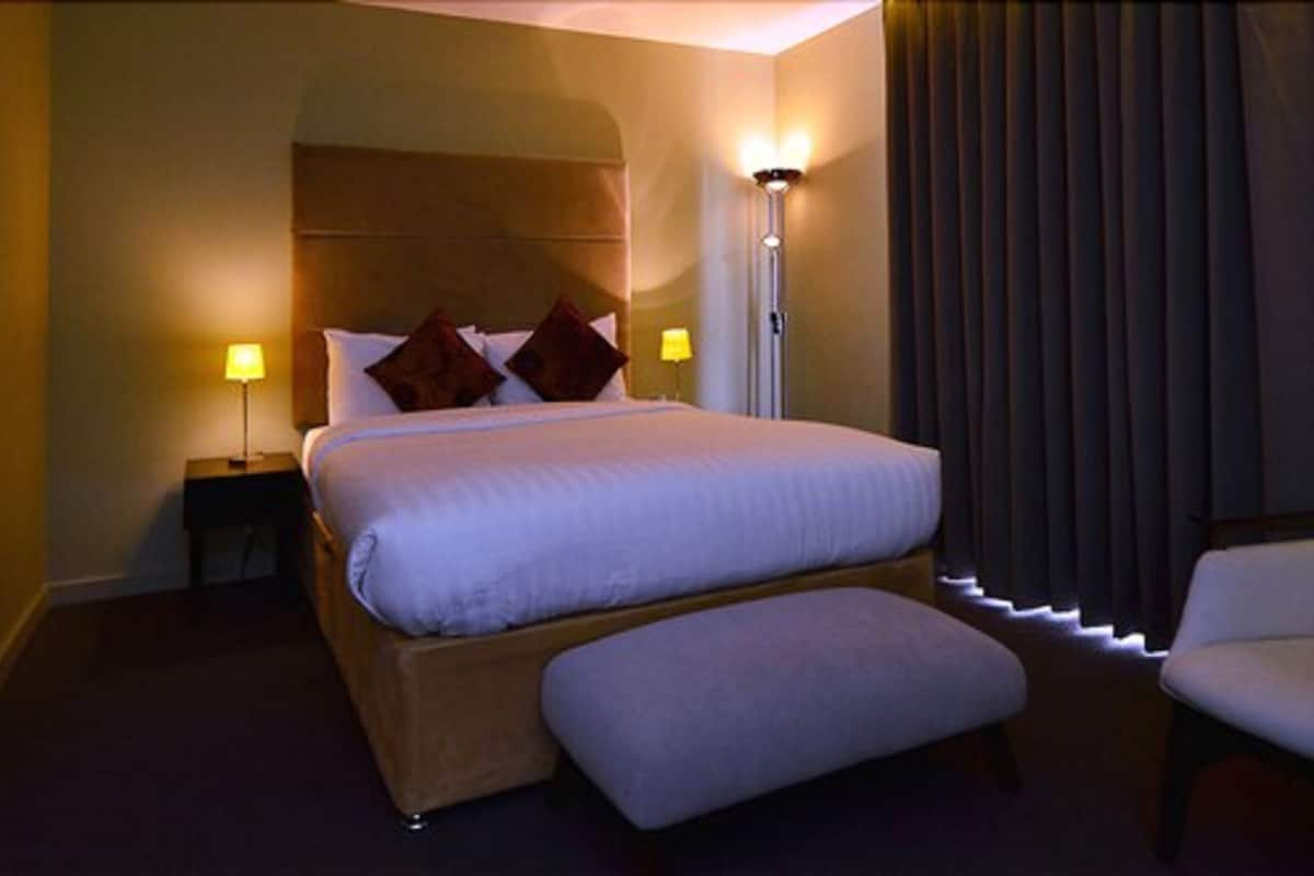 Ein Hotelzimmer mit einem großen Bett, zwei Nachttischen mit Lampen, einer Bank und einem Fenster mit Vorhängen.