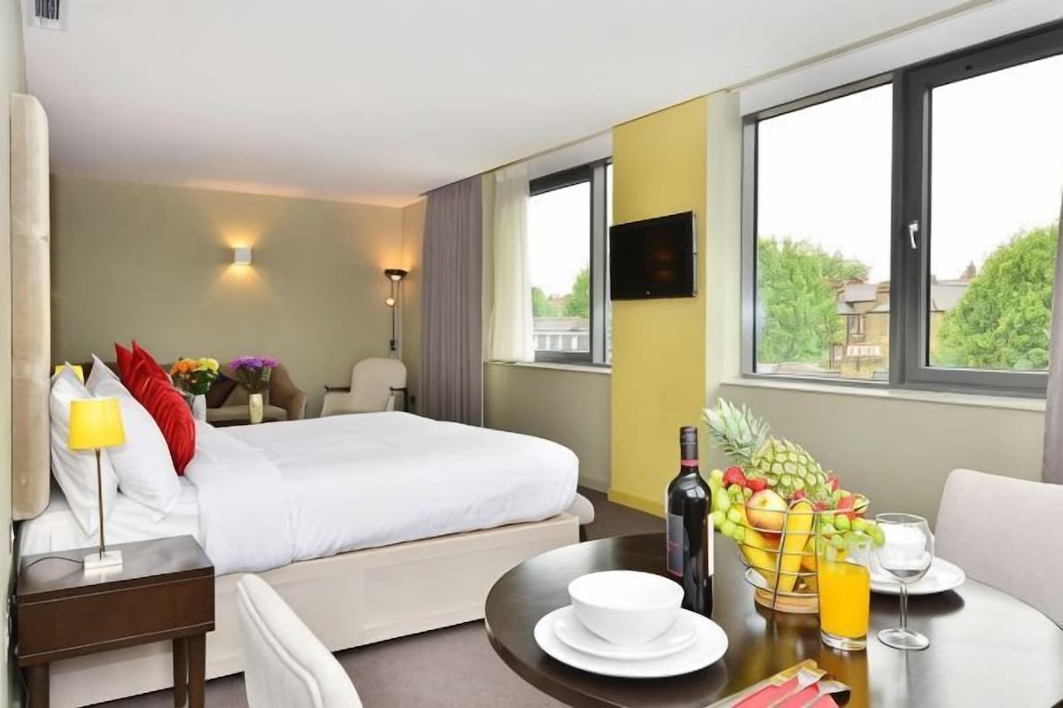Habitación de hotel con cama, mesita de noche, lámpara, silla, mesa de comedor con fruta y bebidas, y televisor.