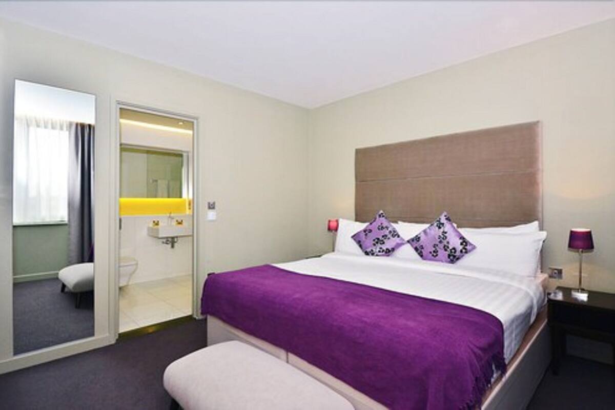 Ein Hotelzimmer mit Bett, Nachttisch, Lampe, Badezimmer mit Spiegel und einer Tür zu einem anderen Raum.
