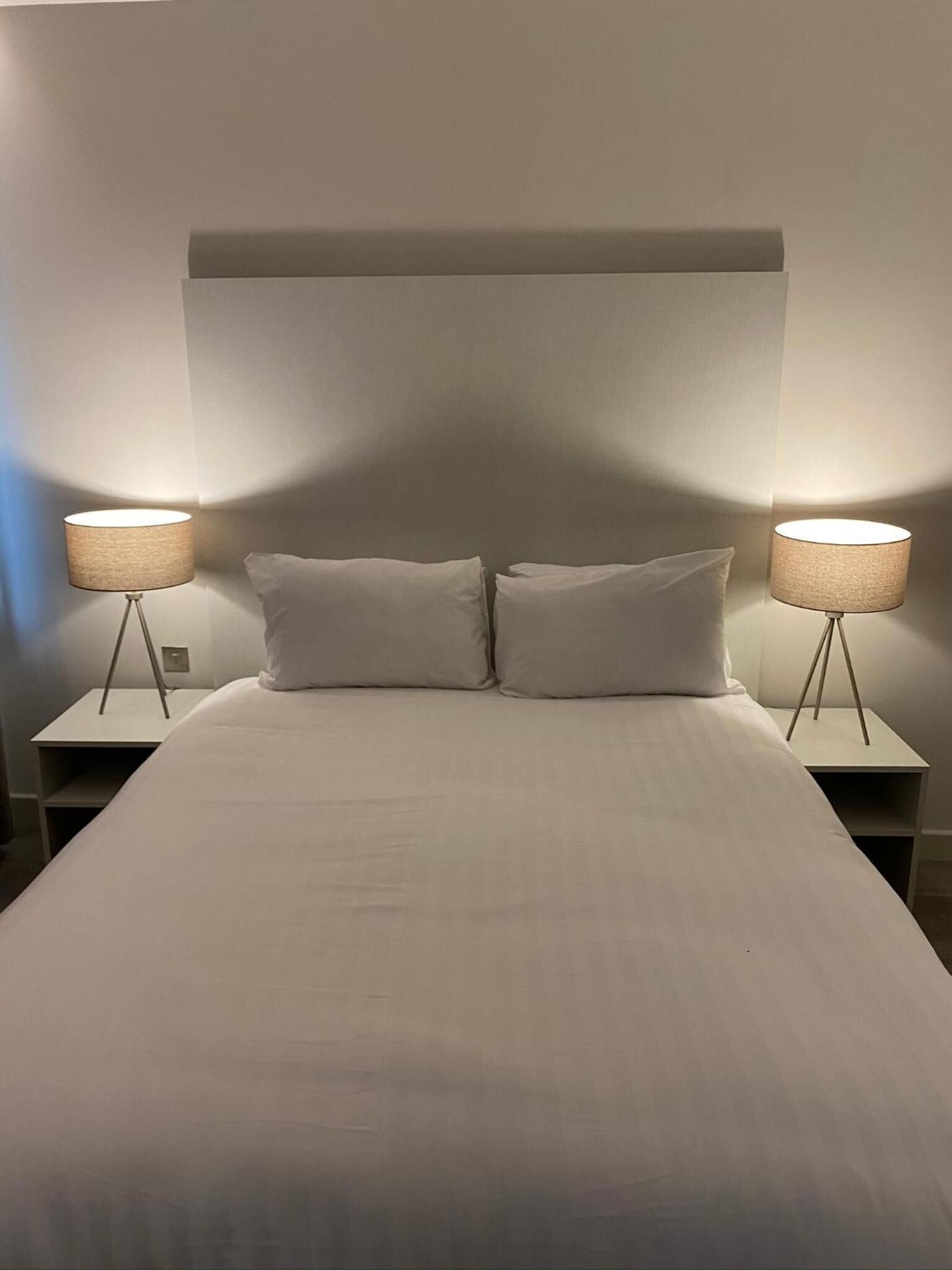 A neatly made bed with two bedside tables, each with a lamp, in a simple room with a plain wall and a sloped ceiling.
