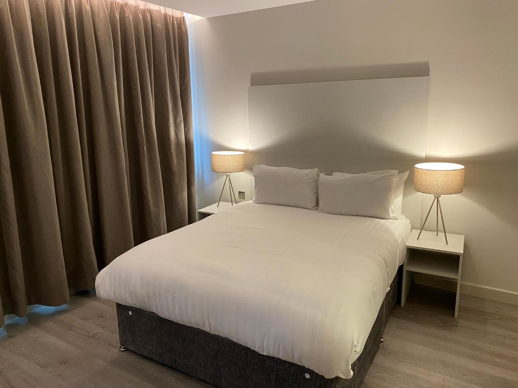 Et moderne hotelværelse med en stor seng, to sengeborde med lamper, en sengegavl og et gardin.
