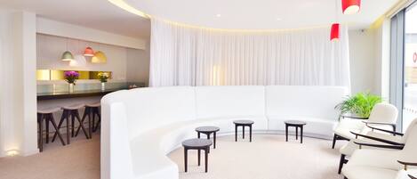 A modern hotel lobby with a curved white seating area, small round tables, and a bar with stools.