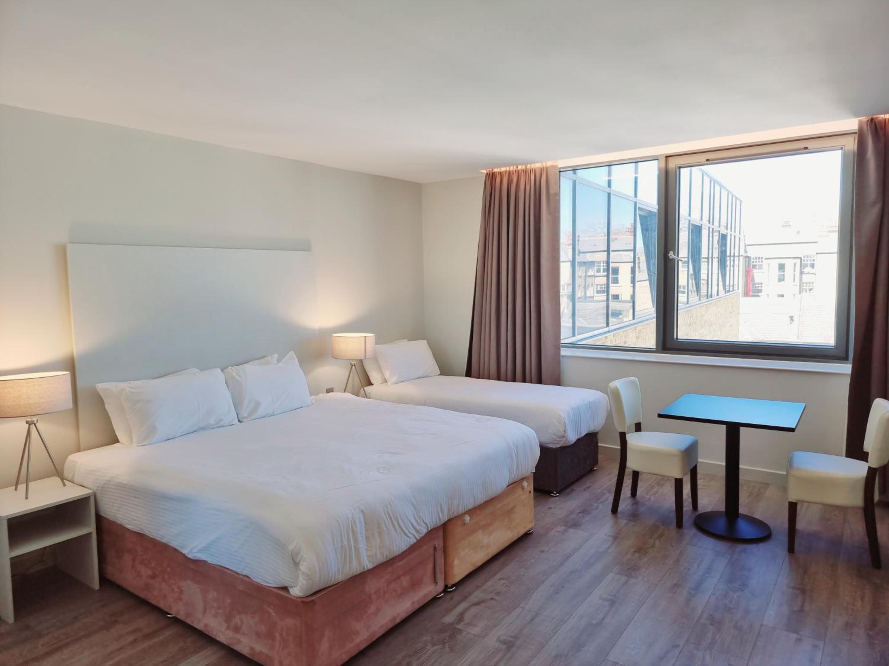 A hotel room with two beds, a small table, two chairs, a lamp, and a window with curtains.