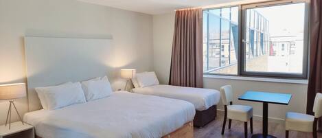 A hotel room with two beds, a small table, two chairs, a lamp, and a window with curtains.