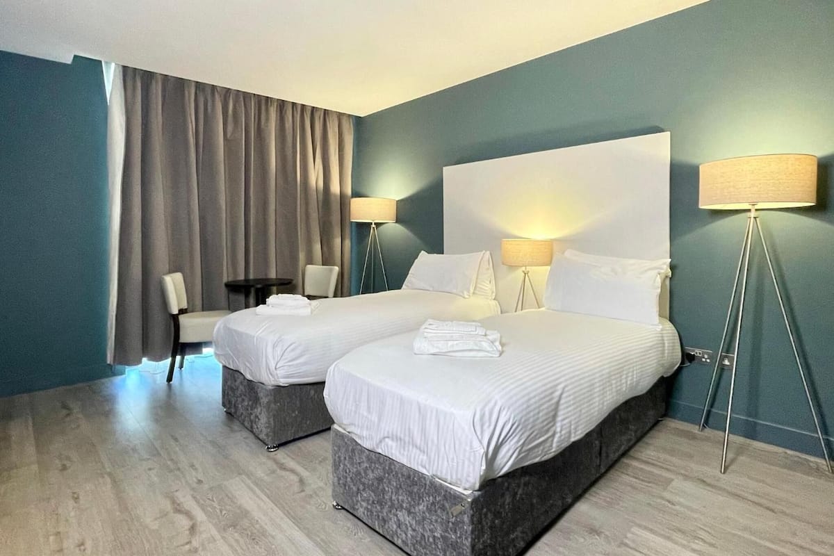 Ein Hotelzimmer mit zwei Betten, einem Sessel, einem Schreibtisch und einer Lampe.