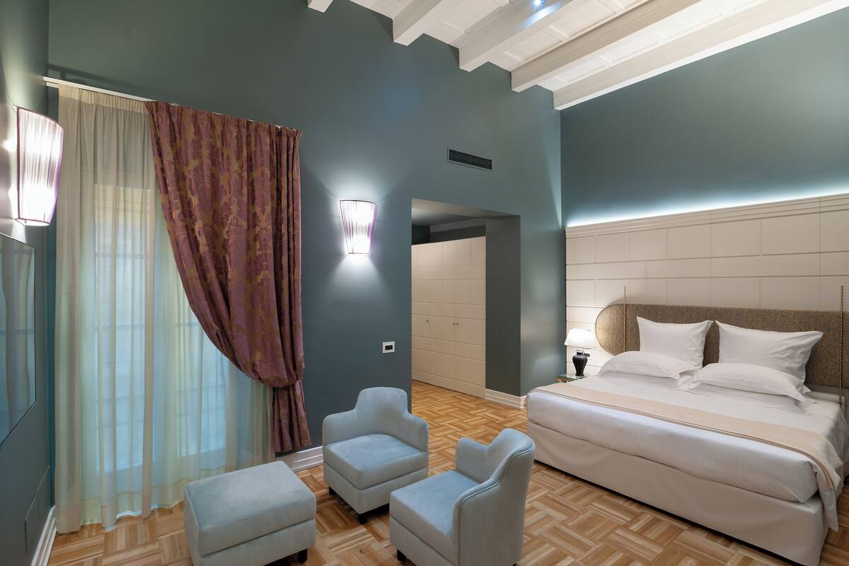 Un dormitorio con cama, dos sillones, una mesita de noche, una lámpara y un ventanal con cortinas traslúcidas y cortina estampada.