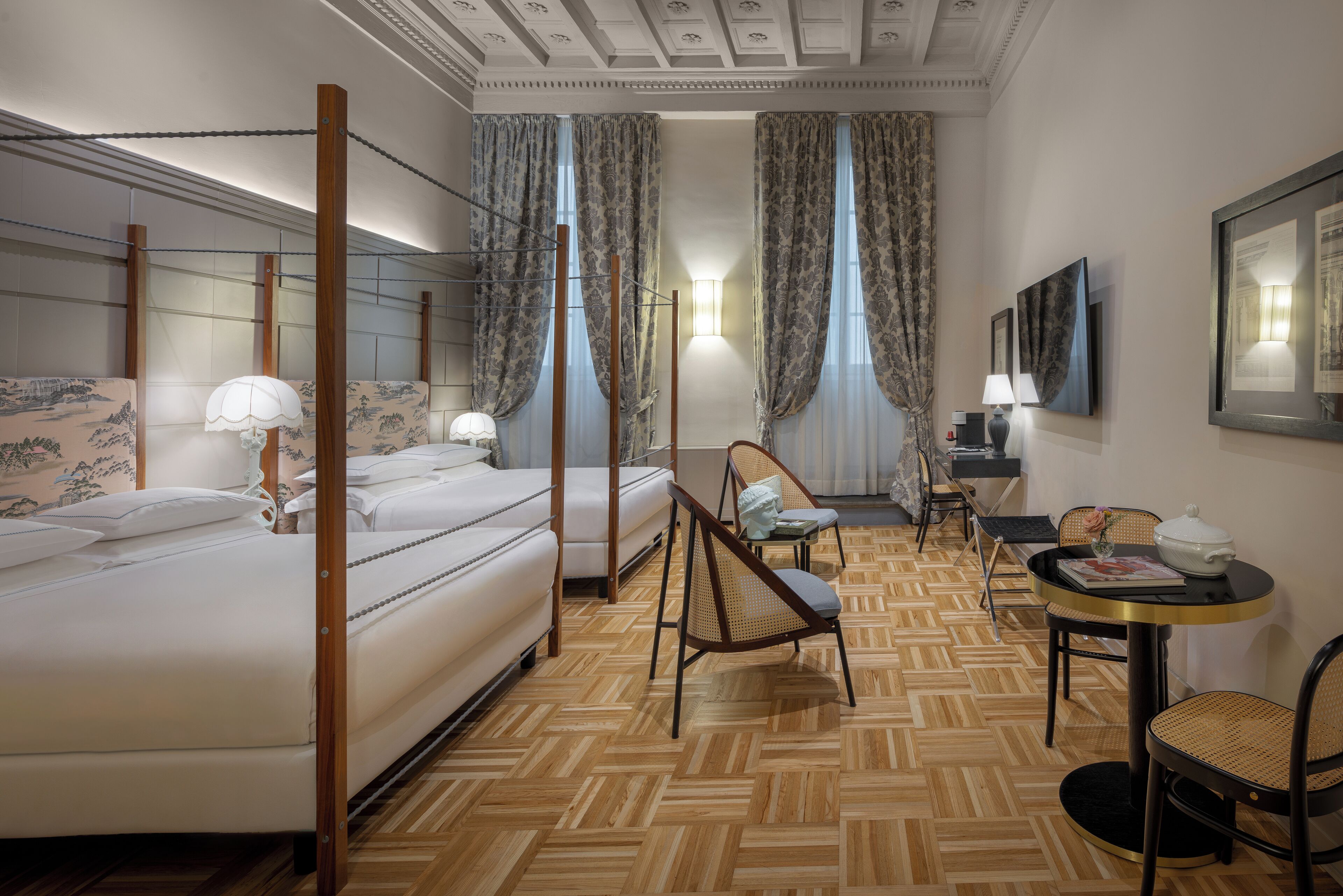 Une chambre d’hôtel avec un lit à baldaquin, un coin salon avec une table et des chaises, et une grande fenêtre avec des rideaux.