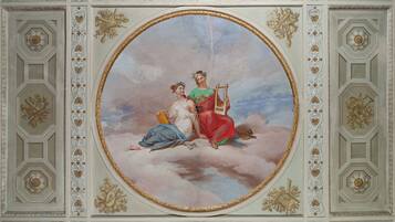 Une fresque ornant le plafond, avec des décorations blanches et dorées très travaillées, représentant deux personnages sur des nuages.