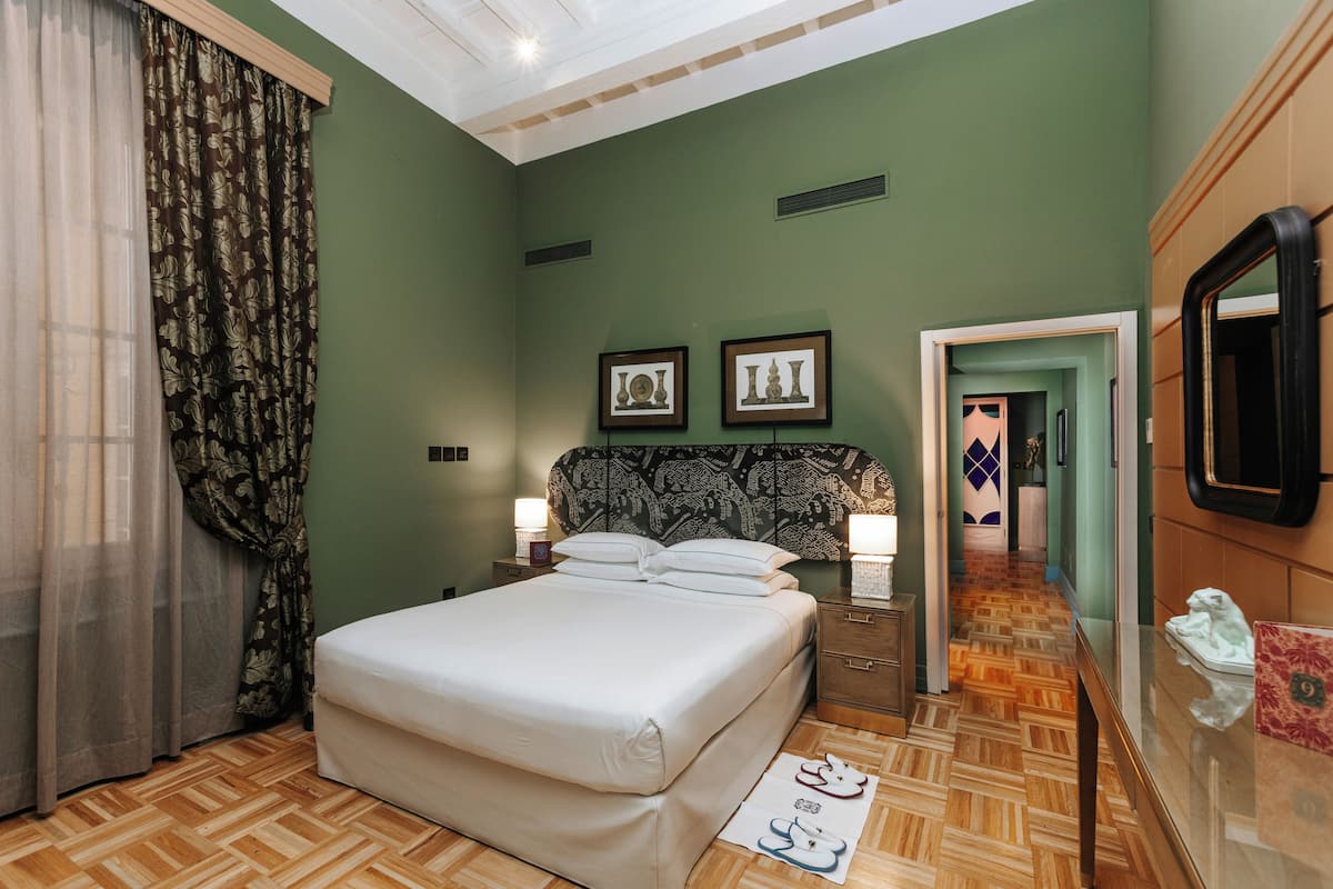 Un dormitorio con cama, mesitas de noche, un espejo y una puerta que da a otra habitación.
