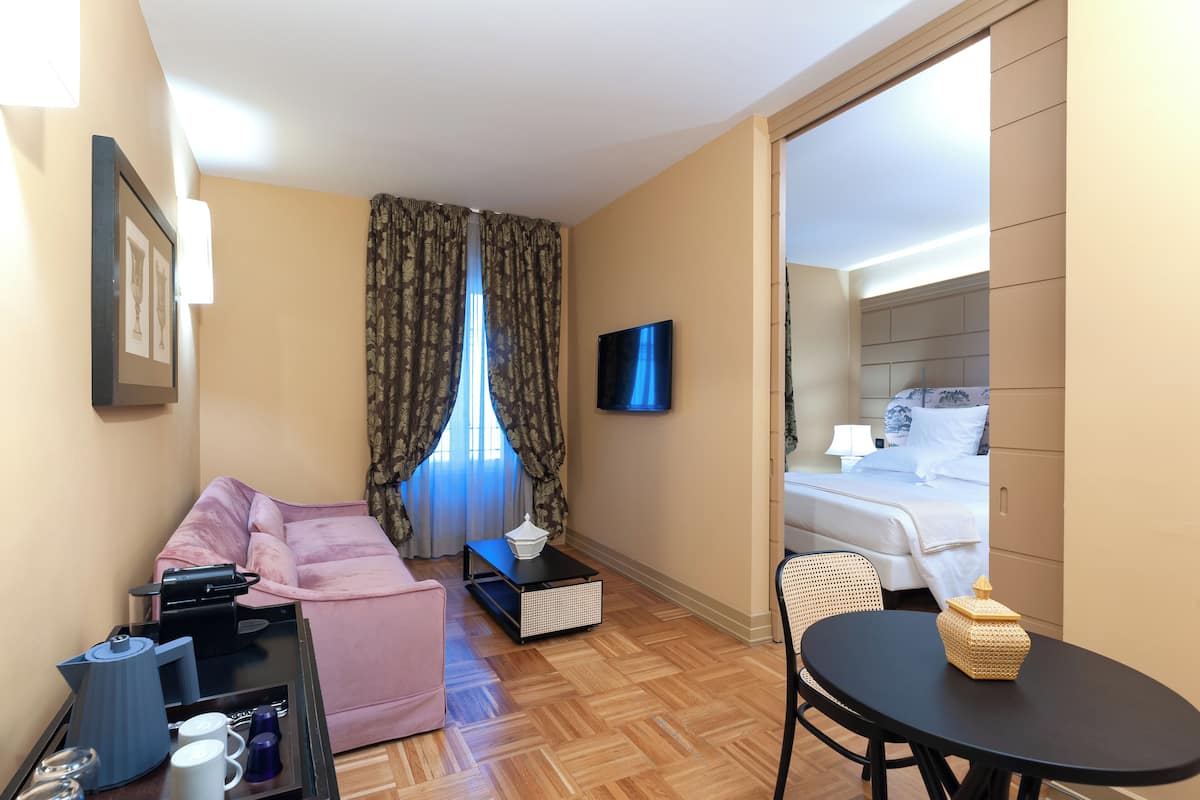 Habitación de hotel con sofá, una mesita con hervidor, televisión, cama con ropa blanca y ventana con cortinas.