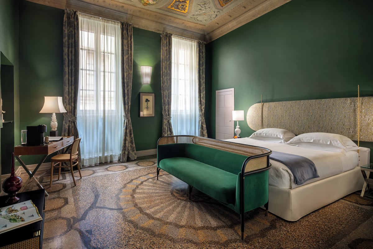 Habitación de hotel con sofá verde, cama con ropa de cama blanca, escritorio con lámpara y ventana grande con cortinas.