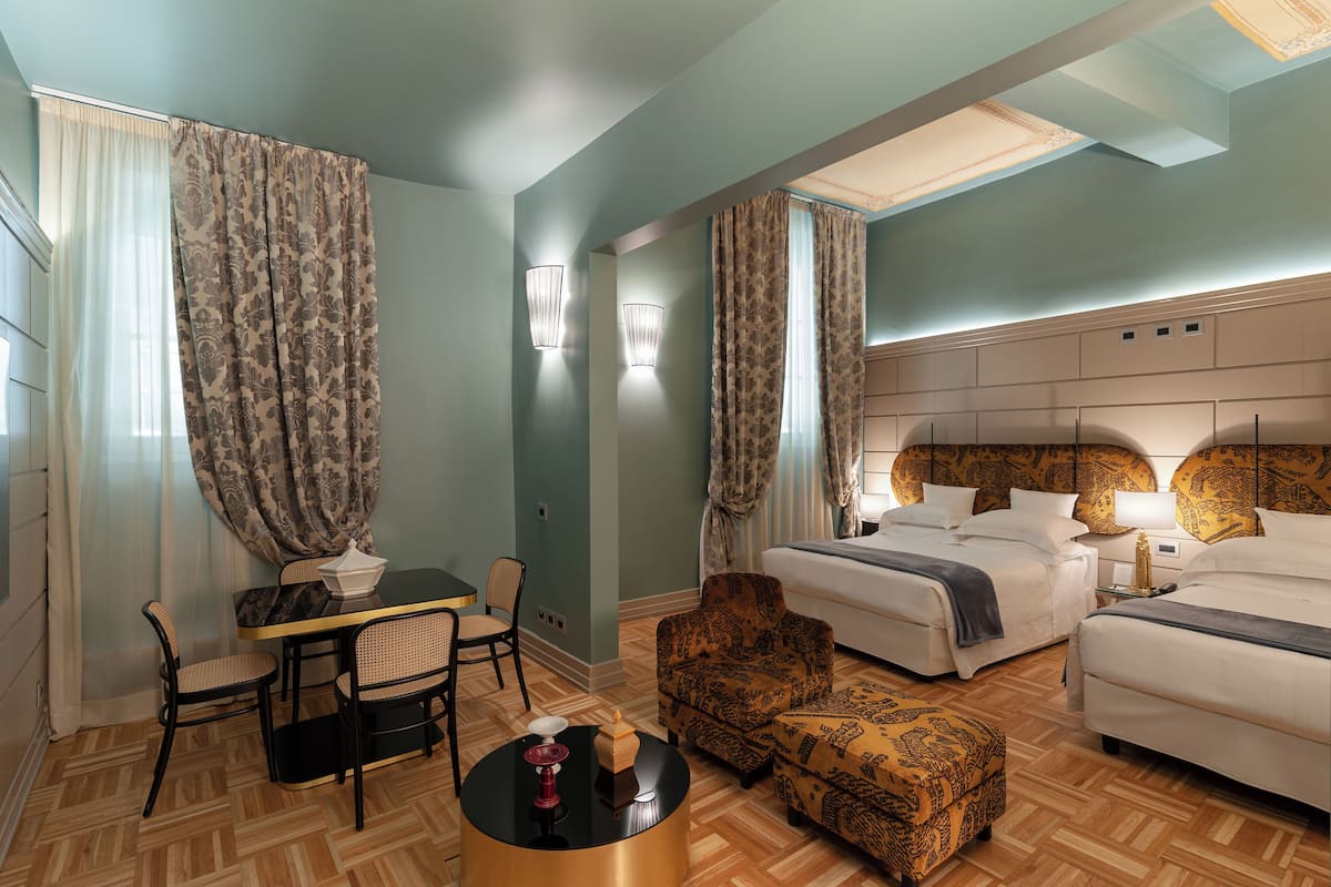 Habitación de hotel con una cama grande, dos camas más pequeñas, una zona de comedor con mesa y sillas, y un sillón estampado.