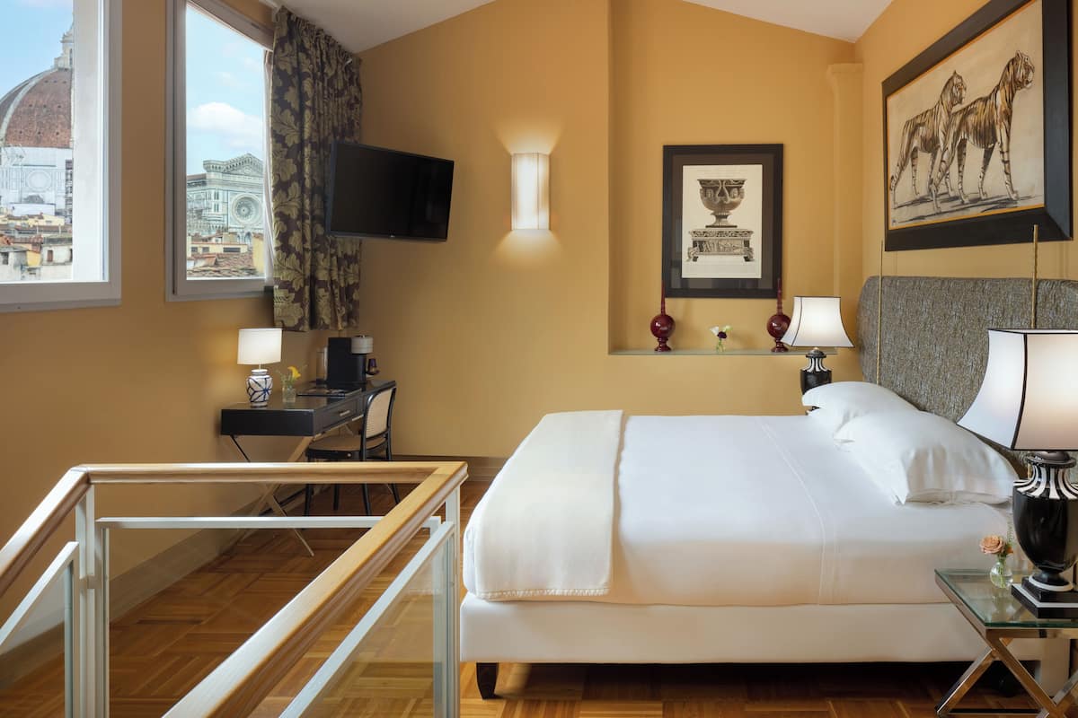 Habitación de hotel con una cama grande, un escritorio con ordenador, un televisor montado en la pared y una ventana con vistas a edificios históricos.