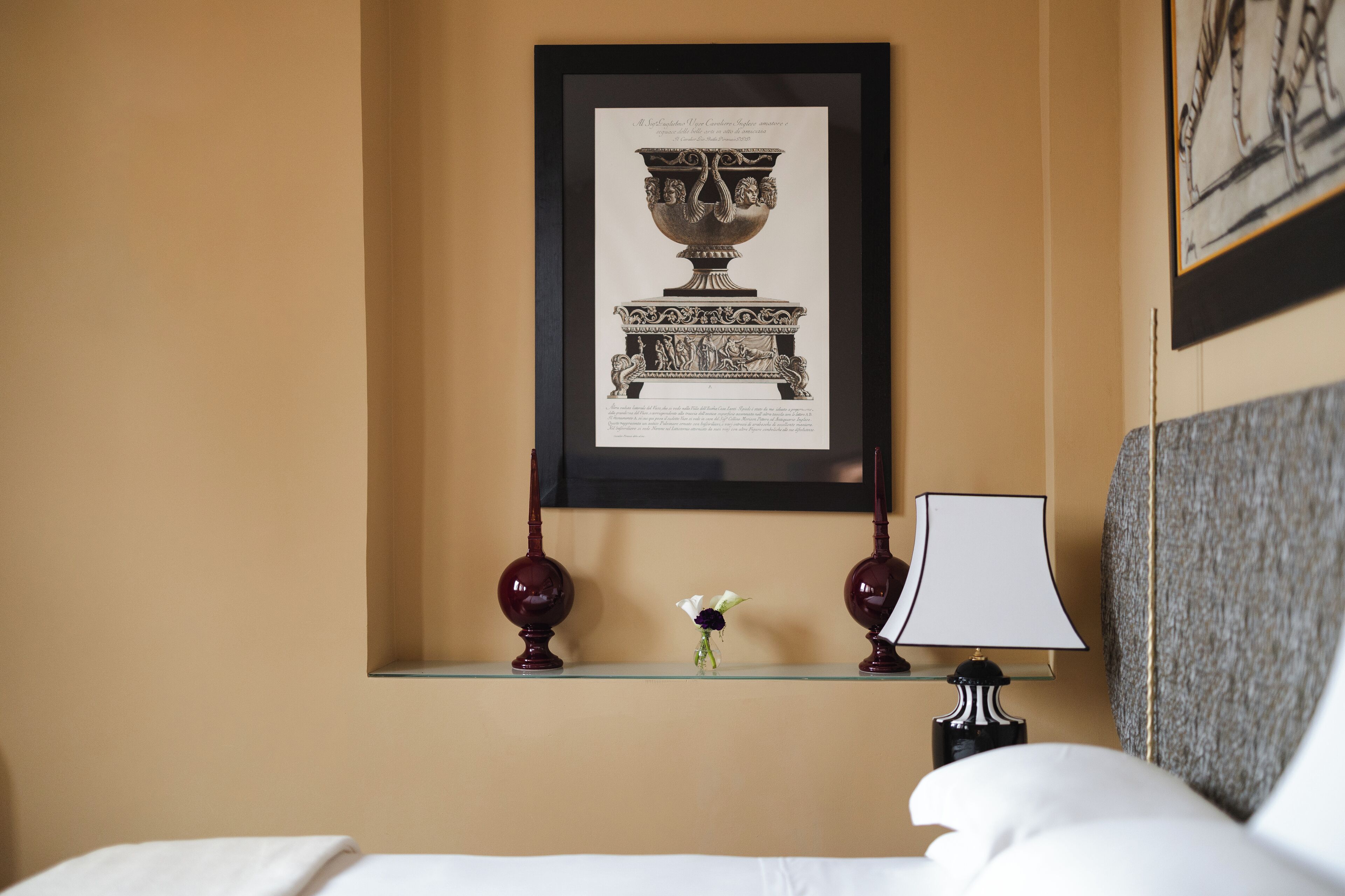 A hotel room with a framed artwork of a classical vase, a small vase with flowers, and a lamp on a bedside table.