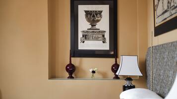 A hotel room with a framed artwork of a classical vase, a small vase with flowers, and a lamp on a bedside table.